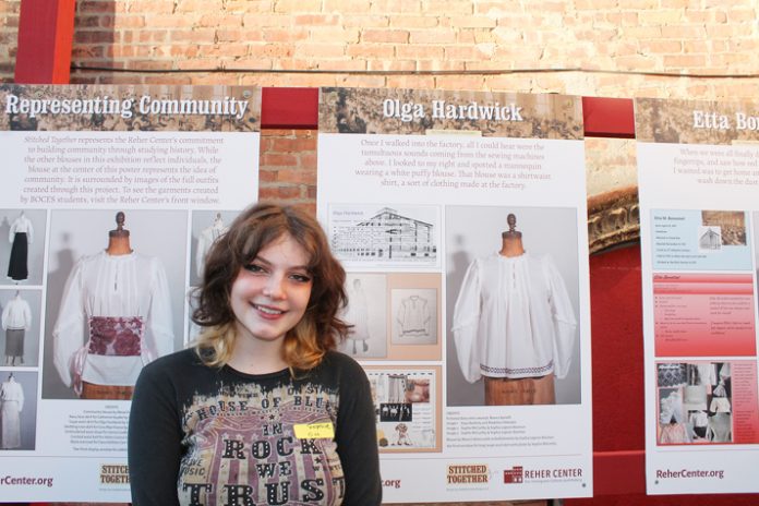 Ulster BOCES Fashion Merchandising & Design student Sophia Legnon-Bozman, from the Rondout Valley Central School District, stands in front of the poster which explains the work behind the creation of an outfit her and fellow students created for Olga Hardwick, a female factory worker who operated a sewing machine at the F. Jacobson and Sons Shirt Factory in Kingston during World War I. The “Stitched Together” project, displayed at the Reher Center for Immigrant History and Culture in Kingston until the end of August, shows the collaboration between Ulster BOCES Fashion Merchandising & Design students, Kingston Catholic High School students, and Rhinebeck High School students, who combined history and imagination to tell the women’s stories.
