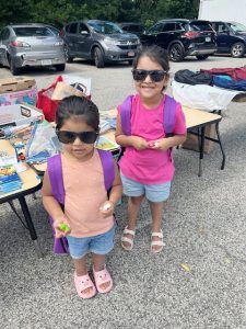 On left, Amirah Solis, age three, who will be entering pre-k, and her sister, Sophia Solis, age five, who will be entering kindergarten, display their new purple knapsacks and other goodies for back-to- school they selected Saturday at the Community Resource Fair.